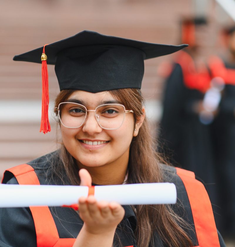 Young indian woman celebrating her achievement, wearing a graduation gown and cap, happily holding her diploma